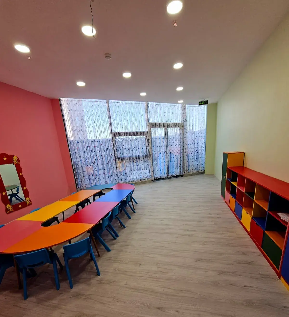 Pembe ve yeşil duvarlı sınıf: mavi sandalyeli uzun masa ve dolaplar
