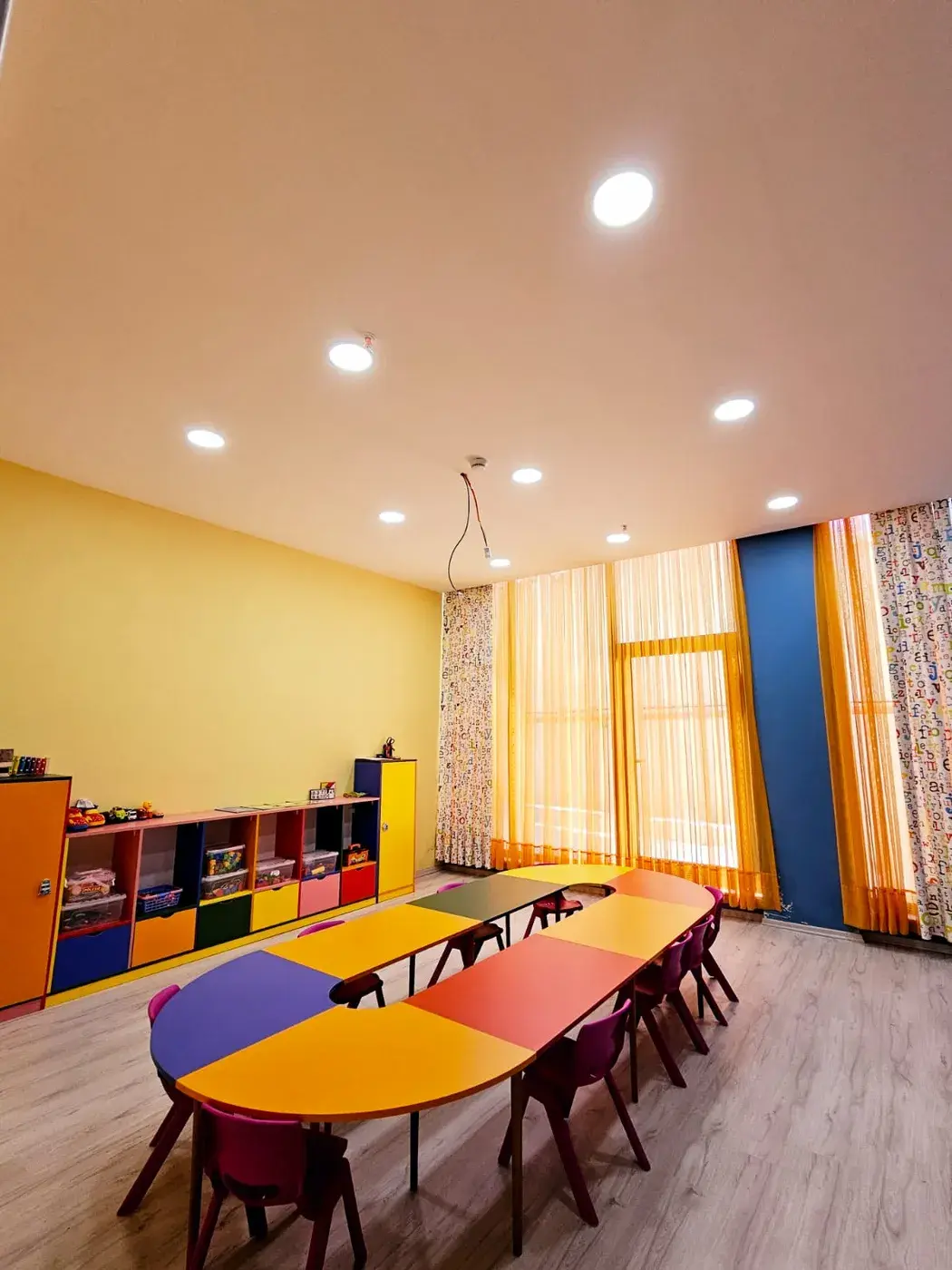 Sarı duvarlı sınıf: renkli oval masa, sandalyeler ve oyuncak rafları